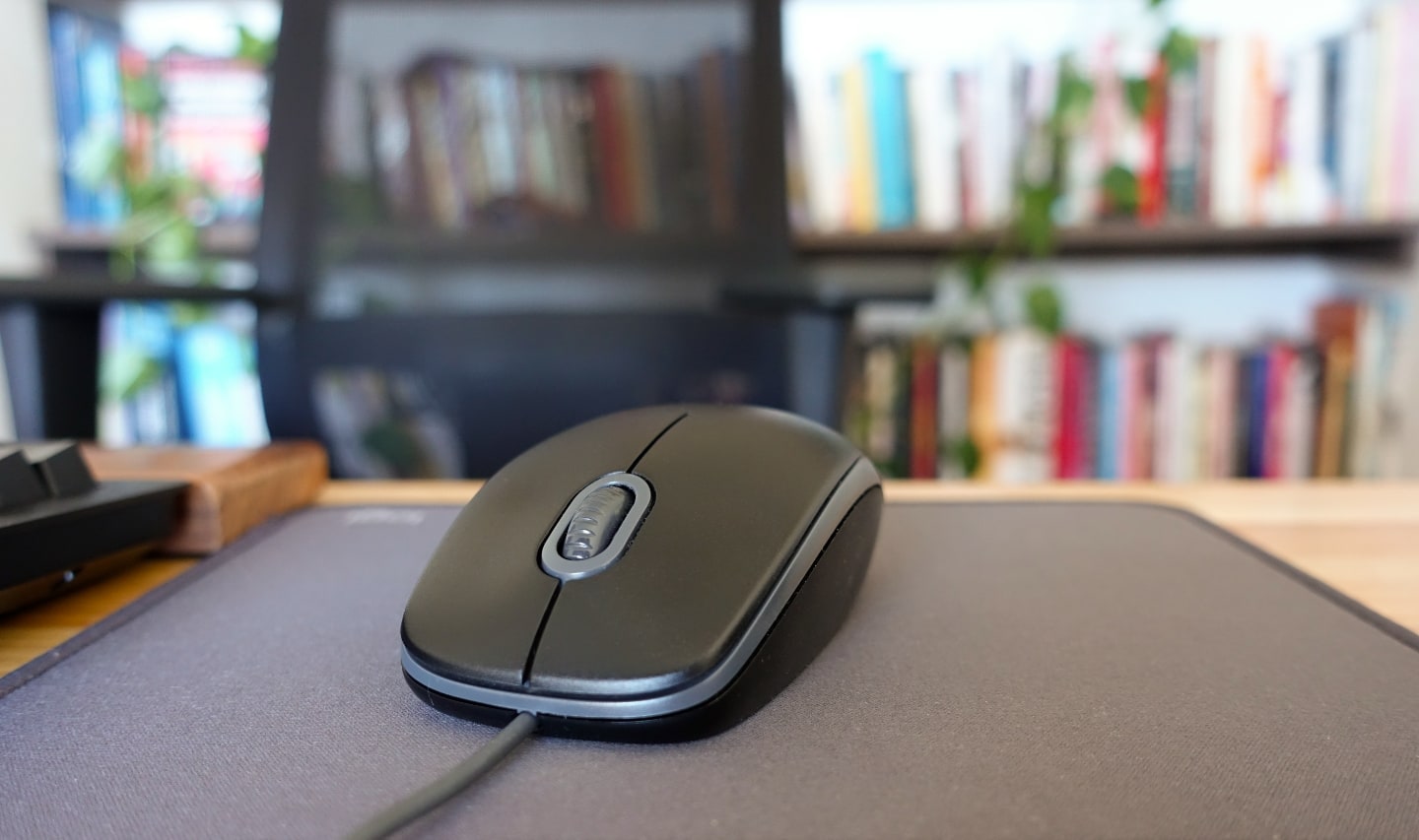 Front photo of a black, wired mouse on a black mousepad on a wooden table. In the blurred background, chair and bookshelf with books.